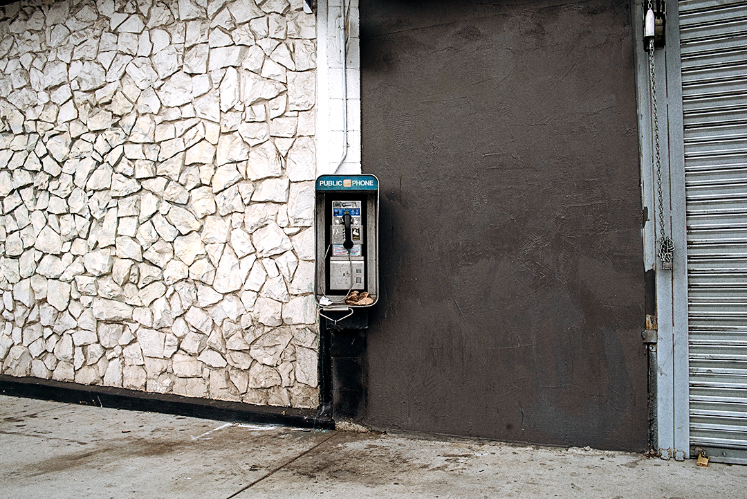 "Street photography of a payphone in front of a connecting rock wall and black wall, urban decay scene in Los Angeles."