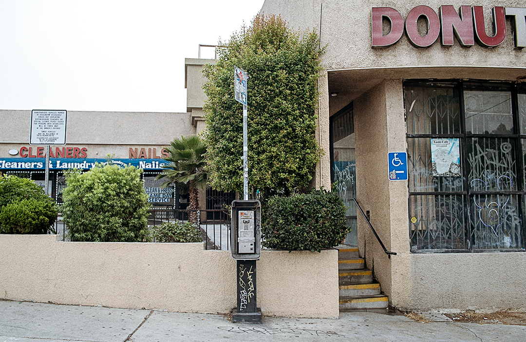 "Street photography of a graffiti-tagged broken payphone in front of a donut shop and plants, urban decay scene in Los Angeles."