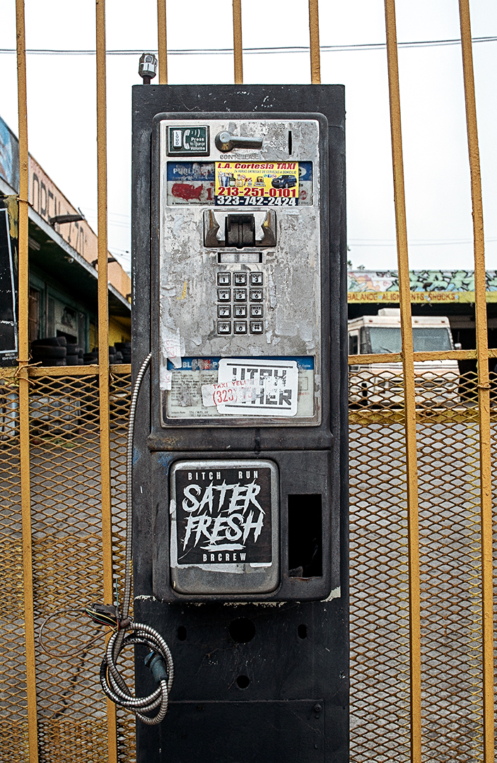Urban decay street photography of an abandoned payphone covered in graffiti and stickers against a yellow fence in Los Angeles.